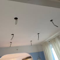 The ceiling features multiple recessed light fixtures with exposed black wiring, reminiscent of recent electrical installations. The room, with its light blue walls and white curtains, is completed by an elegant arched doorway.