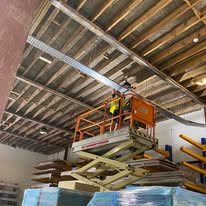An electrician in a yellow vest stands on an orange scissor lift, conducting electrical safety checks while installing or adjusting ductwork on a warehouse ceiling with exposed beams and insulation. Below are stacked materials and shelving units.