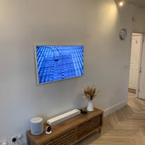 A modern living room features a wall-mounted TV displaying a blue scene, perfect for relaxing after a day of fault finding. Below, a wooden console holds a speaker, small device, and vase of dried flowers. The light wood-patterned floor complements the room's tranquil ambiance, with the door in the background.