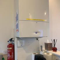 A white wall-mounted boiler is seen in a kitchen, seamlessly blending into the space. Below are paper towels, a tissue box, and a reed diffuser. A red fire extinguisher stands nearby for safety. This setting exemplifies how electrical installations can be both functional and neatly integrated.