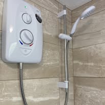 A shower setup features a modern white electric shower unit with an adjustable showerhead on a rail. The unit includes a temperature control dial and hose, ensuring convenience and safety through regular electrical installations checks. Beige tiles elegantly adorn the wall.