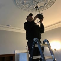 An electrician stands on a ladder, installing a light fixture on a decorative ceiling. The room is softly lit, and the individual is wearing a cap and dark clothing, meticulously working to ensure no fault finding will be necessary later.