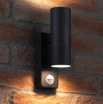 A cylindrical black outdoor wall light, expertly installed by an electrician, is mounted on a brick wall, casting a warm glow downward.