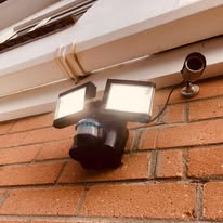 Exterior wall-mounted security system on a brick wall, featuring two bright floodlights and a surveillance camera, expertly installed under the eaves for optimal electrical safety checks.