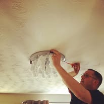 A person standing on a ladder is installing or fixing a ceiling light fixture. With a screwdriver in hand, they carefully adjust the fixture, ensuring electrical installations are secure. The room's softly lit ambiance highlights the ceiling's textured pattern.
