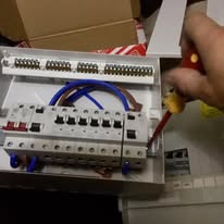 A person's hand deftly uses a screwdriver on an open electrical panel, conducting essential electrical safety checks. The panel reveals several circuit breakers and tangled wires, while in the background, a partially visible box and scattered papers complete the scene.