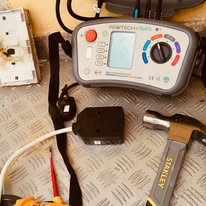A multimeter with various settings and a display rests on a textured metal surface. Nearby, a detached electrical outlet, hammer, and other tools hint at electrical safety checks or consumer unit upgrade work in progress. The scene suggests meticulous attention to electrical maintenance.