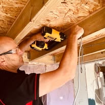 A person wearing safety glasses uses a power drill on wooden beams at a construction site, indicating renovation or building work. Wiring is visible nearby, suggesting potential electrical installations. The setting appears to be indoors.