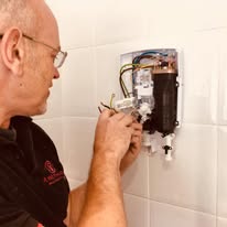 A short-haired electrician in a black shirt skillfully tackles fault finding, installing or repairing an electrical device against a white tiled wall, expertly managing several wires.