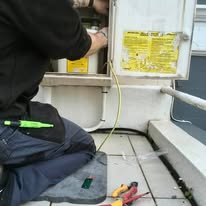 An electrician is kneeling on a rooftop, working diligently on an electrical box. Various tools are scattered around, including pliers and a utility knife. A yellow cable runs from the box. Dressed in a dark shirt and jeans, they focus intently on ensuring all electrical installations are precise and safe.
