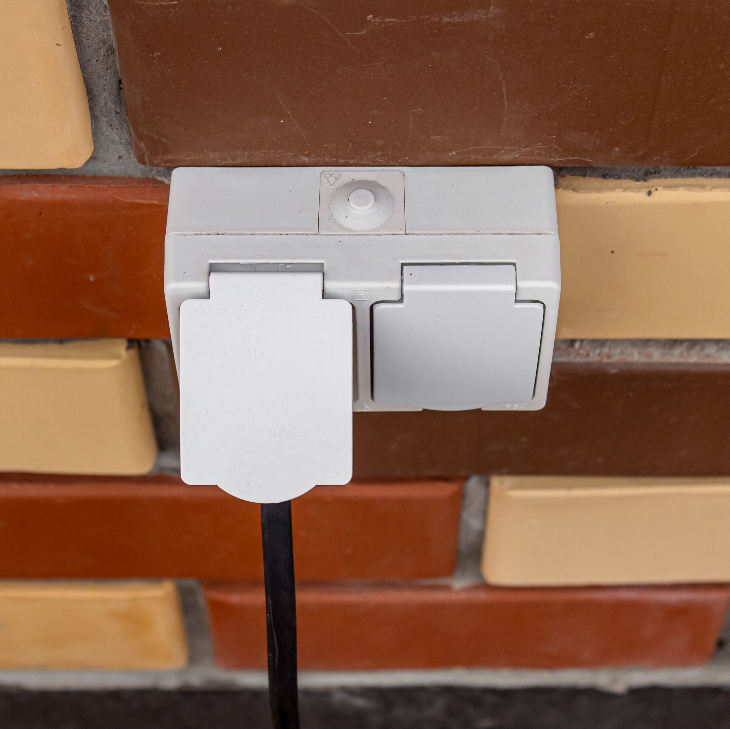 A double white electrical outlet, expertly installed as part of comprehensive electrical installations, features a black power cord in the left socket. It is mounted on a brick wall with brown and beige bricks, while the right socket remains safely covered with a protective flap.