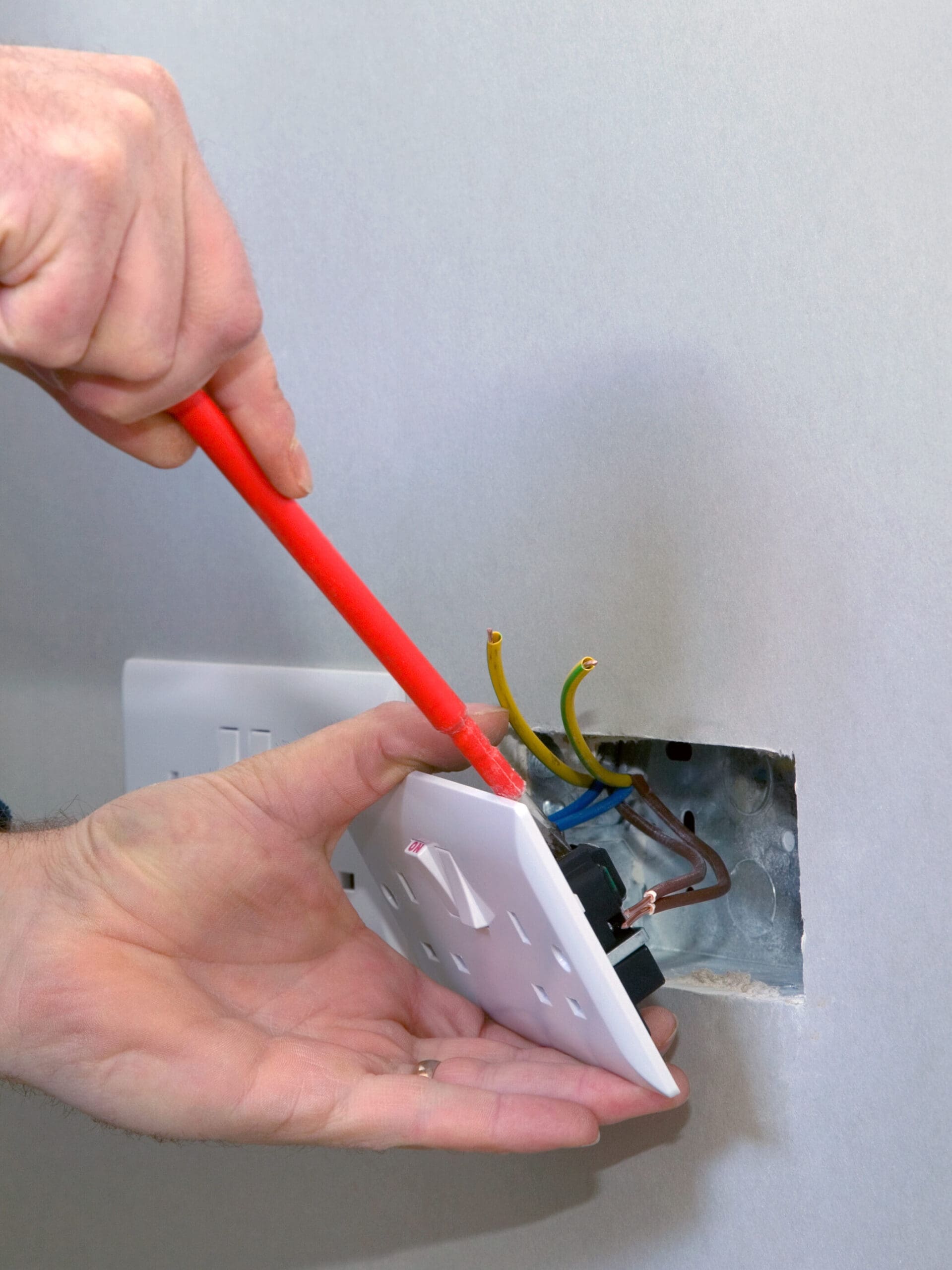 A person carefully uses a red screwdriver for electrical installations, either installing or removing a white outlet cover from the wall. Several exposed wires are visible, with a hand supporting the cover to ensure precision and safety during the process.