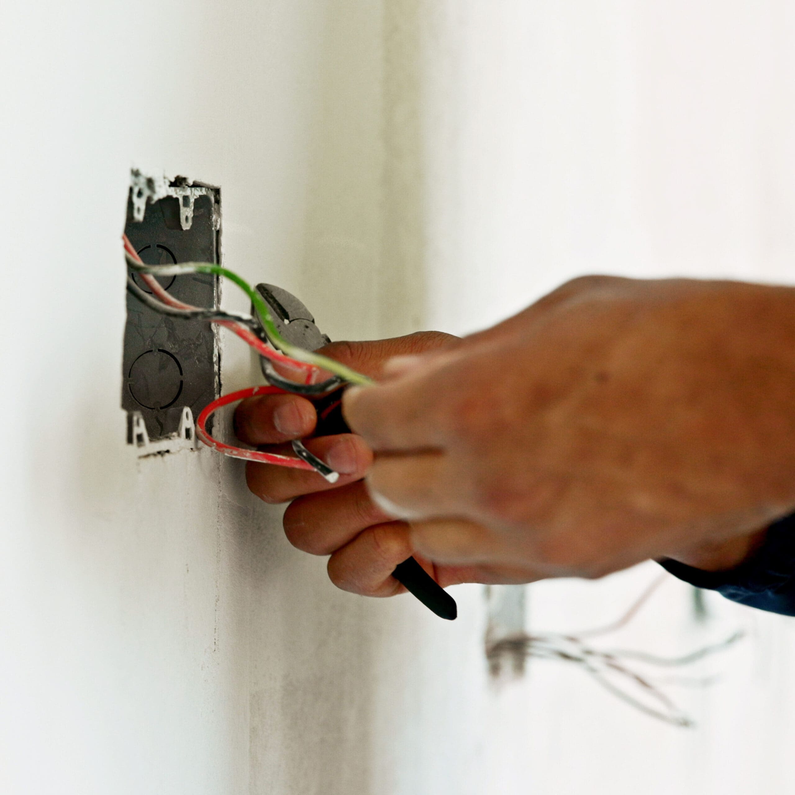 An electrician skillfully uses pliers to work on colorful electrical wires protruding from a wall socket. The area around the socket suggests ongoing construction or repair, with a focus on upgrading the consumer unit for enhanced safety. The person's hand remains sharply in focus.