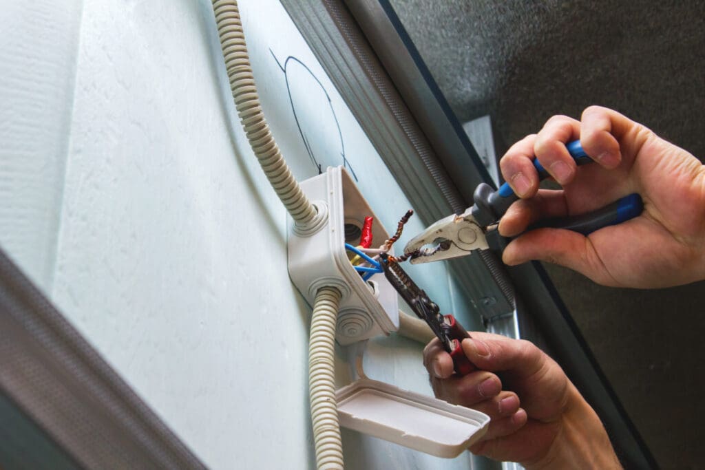 A person is using pliers to work on electrical wires inside a white junction box on a wall, conducting essential electrical safety checks. The box cover is open, and wires are visible. The scene is well-lit, suggesting an indoor setting focused on precision and safety.