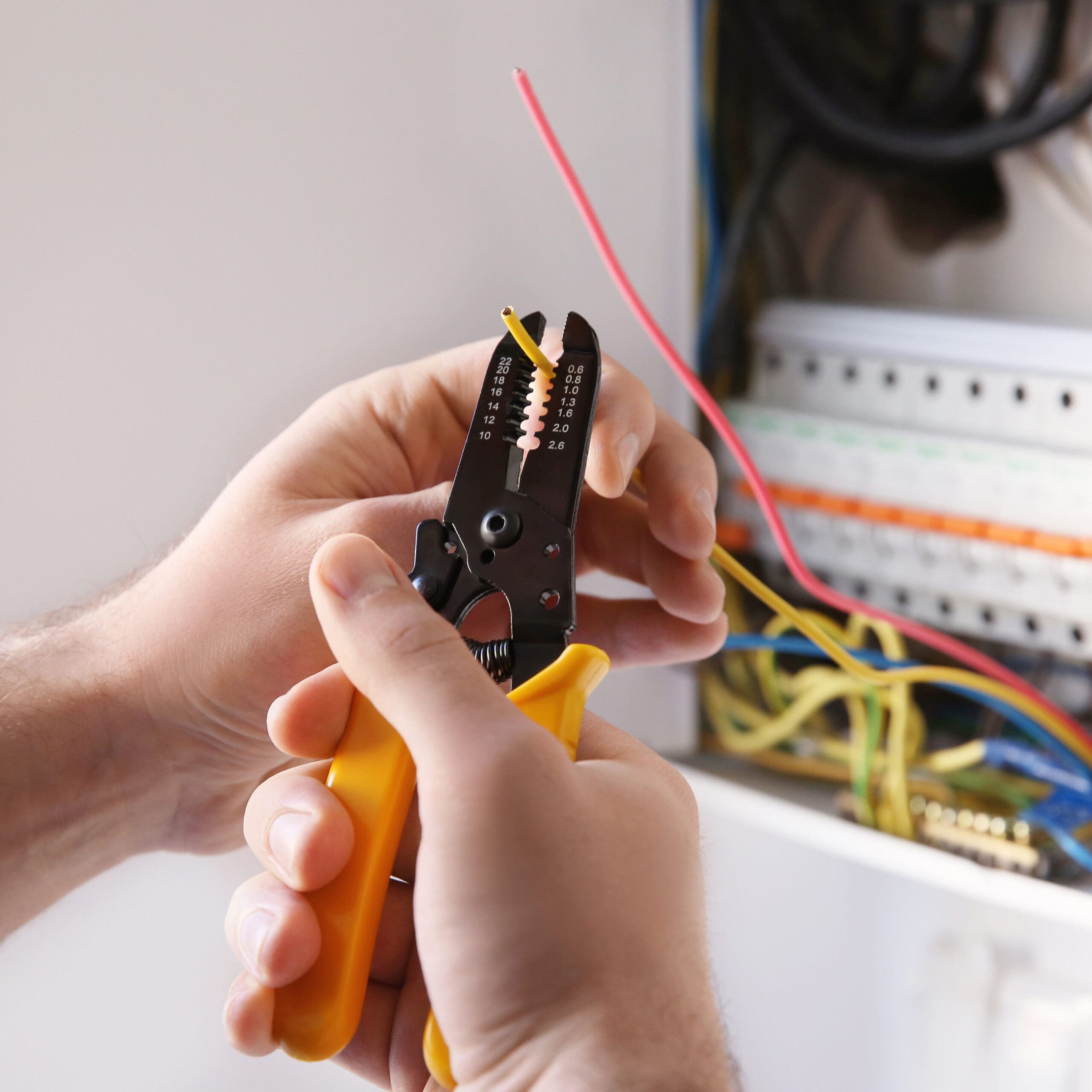 An electrician skillfully uses wire strippers with yellow handles to remove insulation from a yellow wire near an electrical panel filled with multiple colored wires, demonstrating expertise in electrical installations.