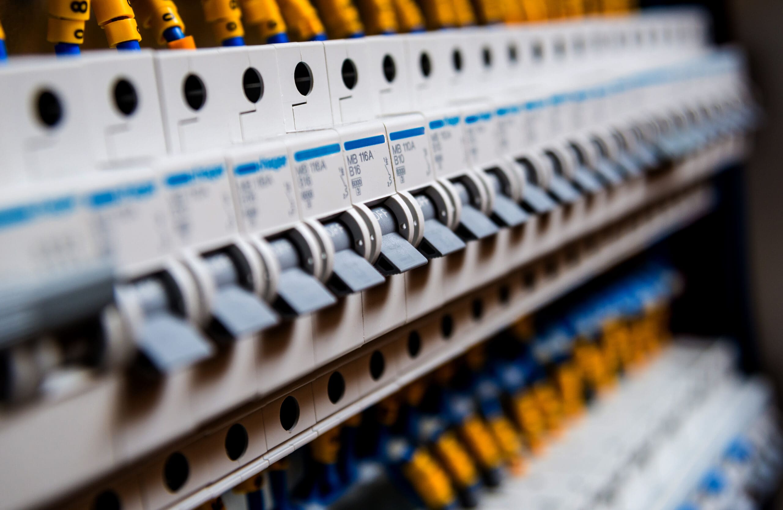 Close-up of electrical circuit breakers lined up in a row. Each breaker has a black switch and is labeled with text. Yellow and blue wire connectors create a colorful pattern on top—an essential setup for any electrician conducting electrical safety checks.