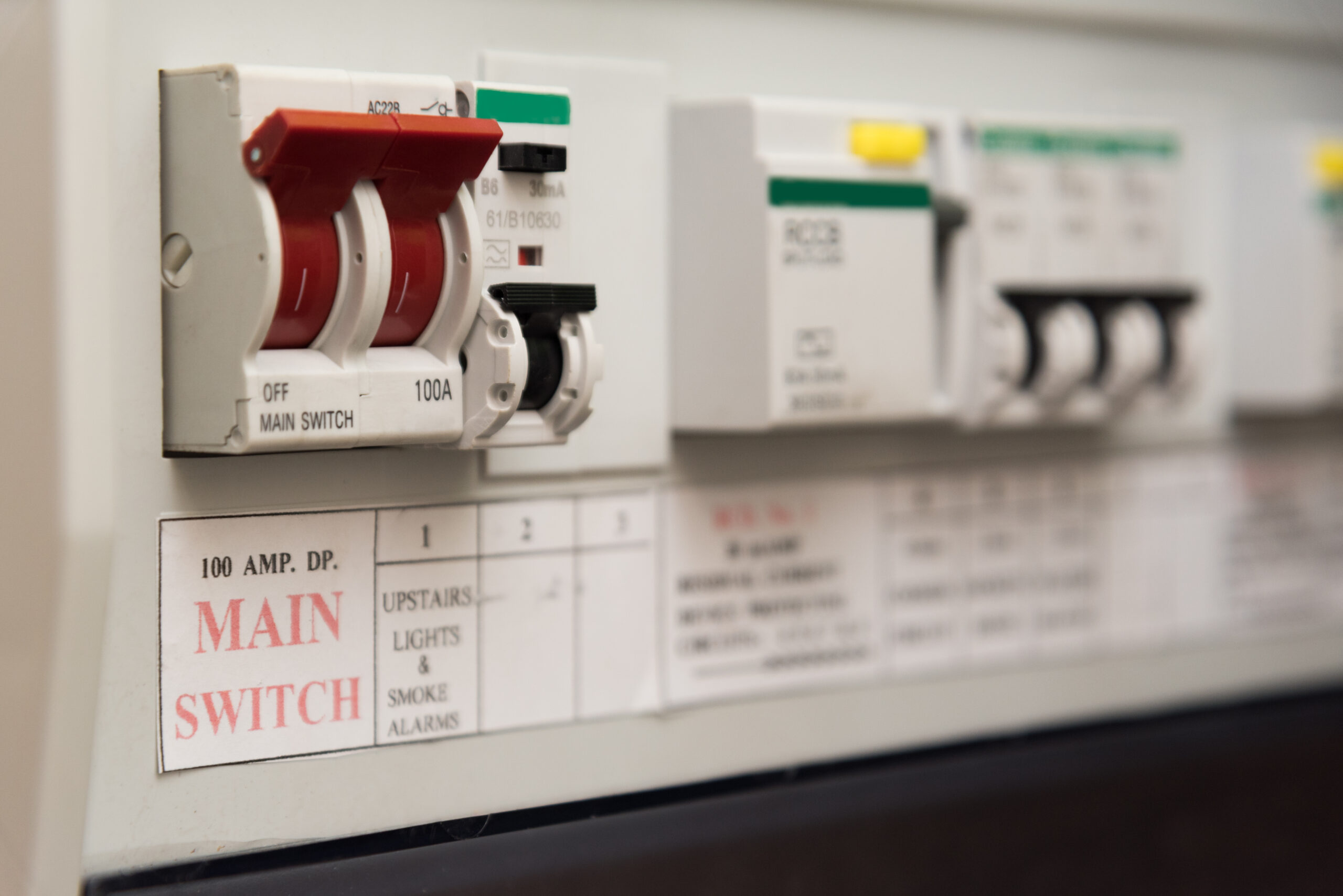 Close-up of an electrical panel with a large red main switch labeled "100 AMP. DP. MAIN SWITCH" alongside smaller switches and circuit breakers. Labels indicate functions like "Upstairs Lights & Smoke Alarms," illustrating the importance of regular electrical safety checks for optimal performance.