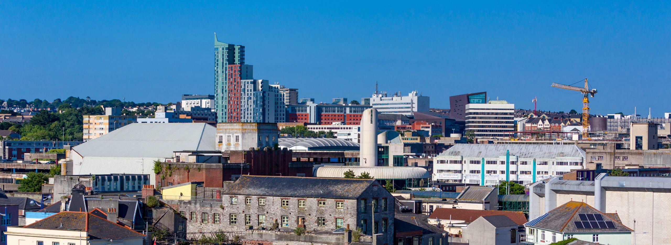Panoramic view of an urban skyline featuring a blend of modern high-rises and older buildings under a clear blue sky. A construction crane is visible on the right, highlighting ongoing development, possibly for new electrical installations.
