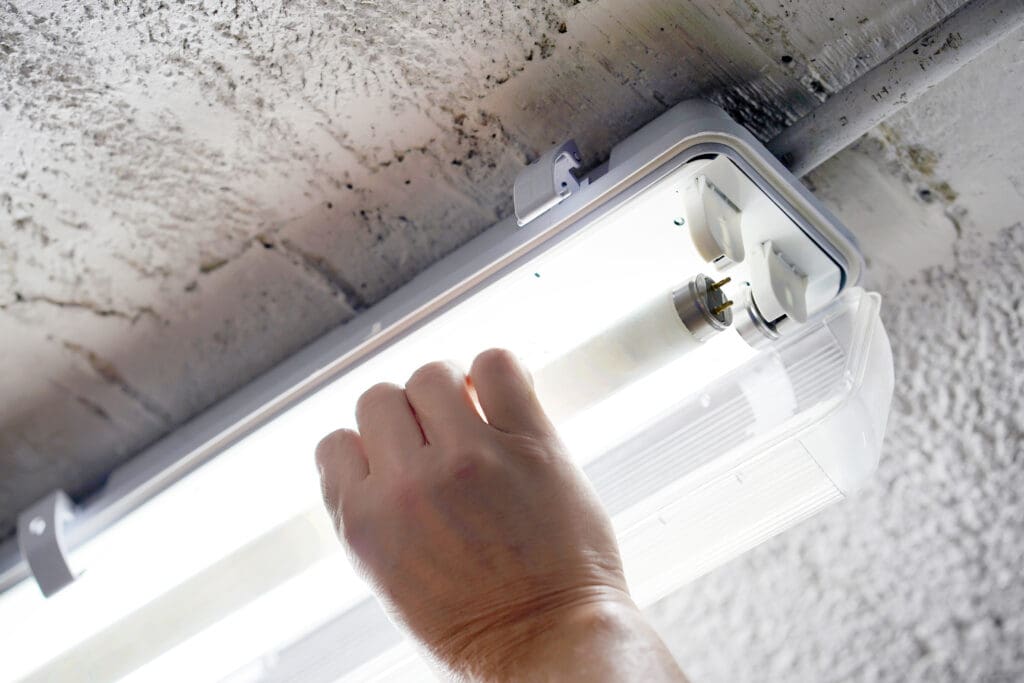 An electrician's hand deftly installs or removes a fluorescent tube light in a ceiling fixture. The light is switched on, casting a bright glow, as part of the ongoing electrical installations.