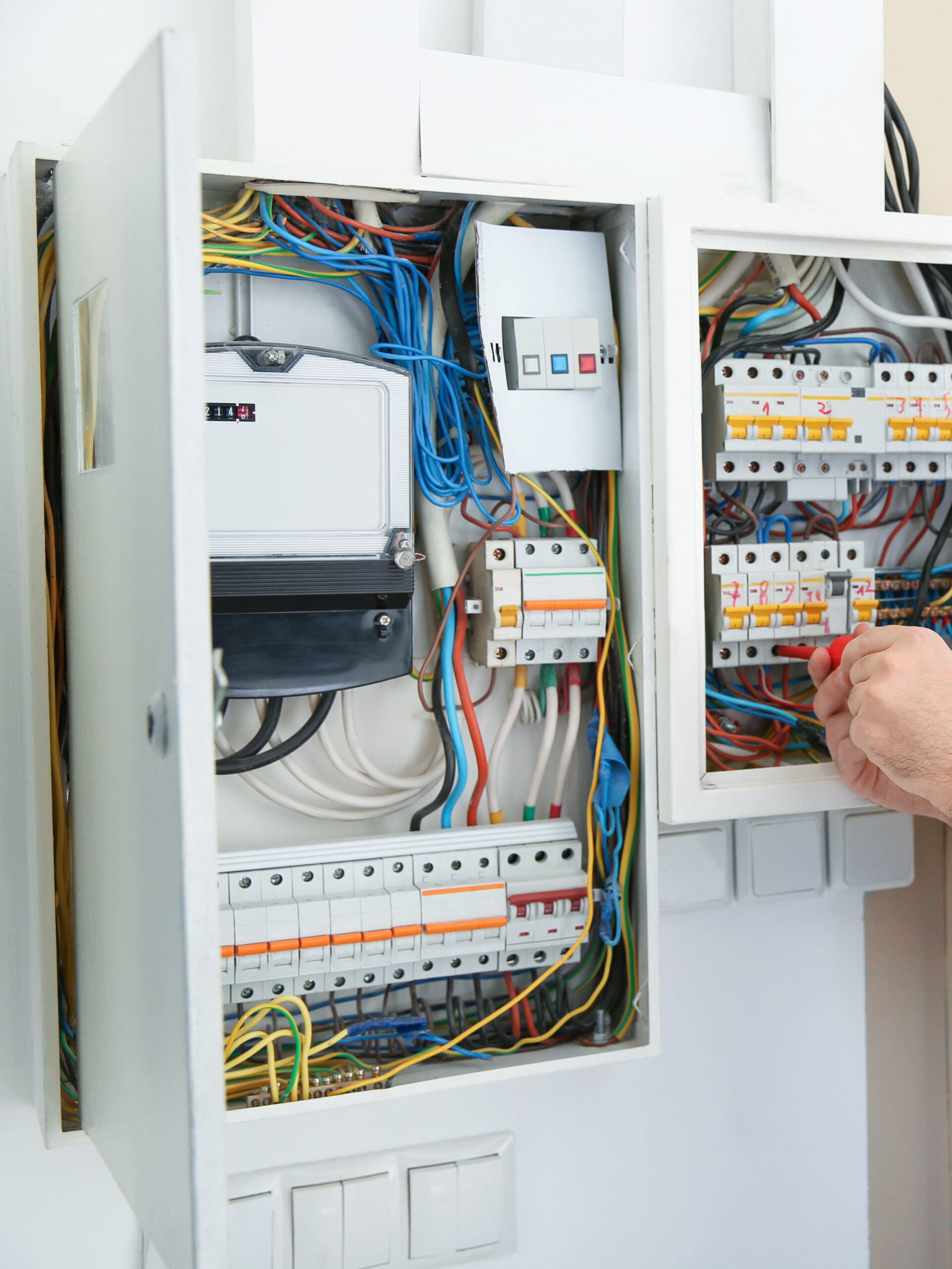 A person adjusts wires in an open electrical panel, skillfully conducting electrical safety checks. The colorful wiring and circuit breakers remain exposed, with a meter nearby. The panels are wall-mounted, as the person's hand is visible steadying a tool near the breakers for fault finding.