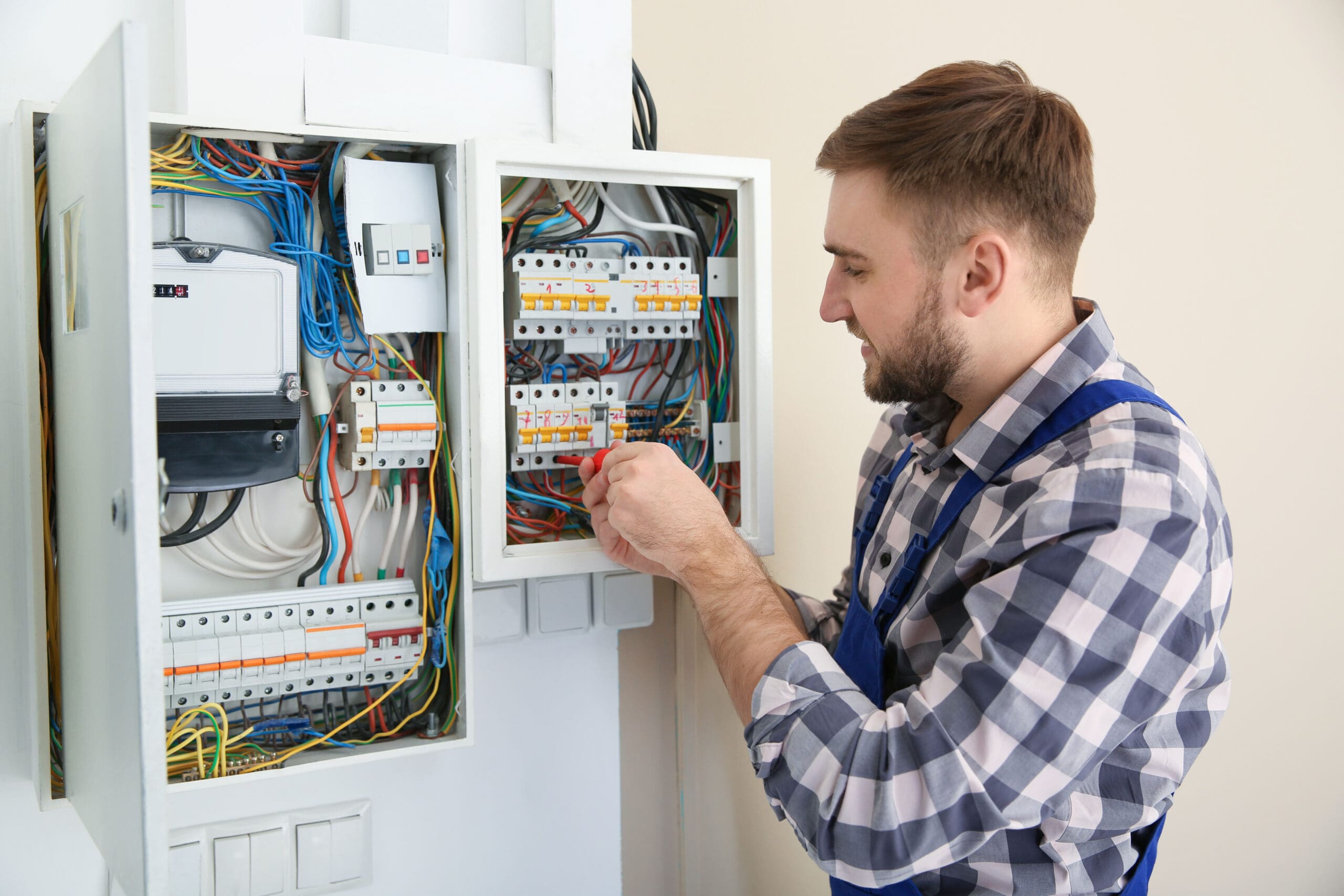 An electrician in a checkered shirt and blue overalls is adjusting wires in an open electrical circuit breaker panel, perhaps for a consumer unit upgrade. Various colorful cables are visible against the light-colored walls.