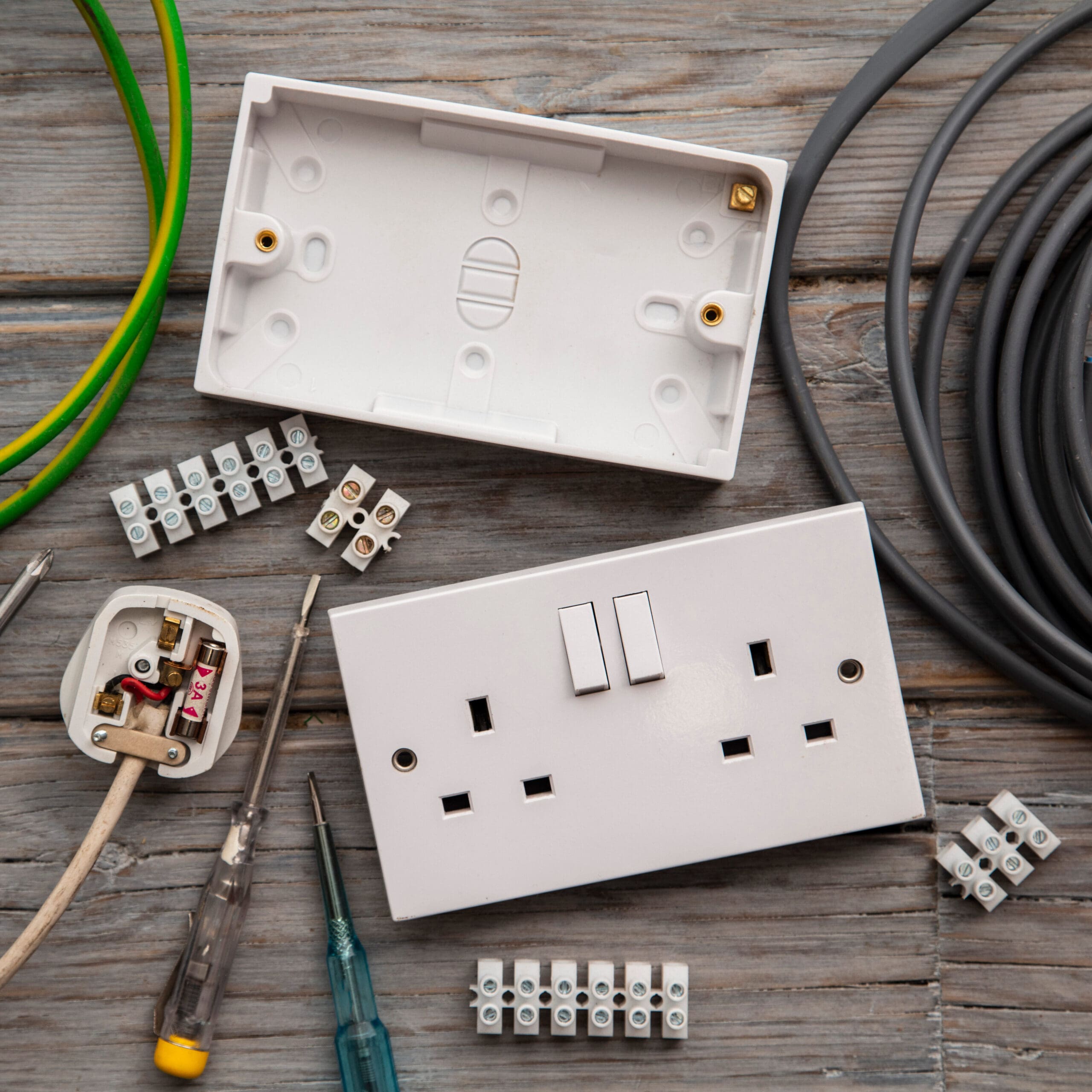 Tools and components for electrical work on a wooden surface, perfect for an electrician, including a UK-style double plug socket, wiring, terminal connectors, two screwdrivers, and a white faceplate—ideal for conducting thorough electrical safety checks.