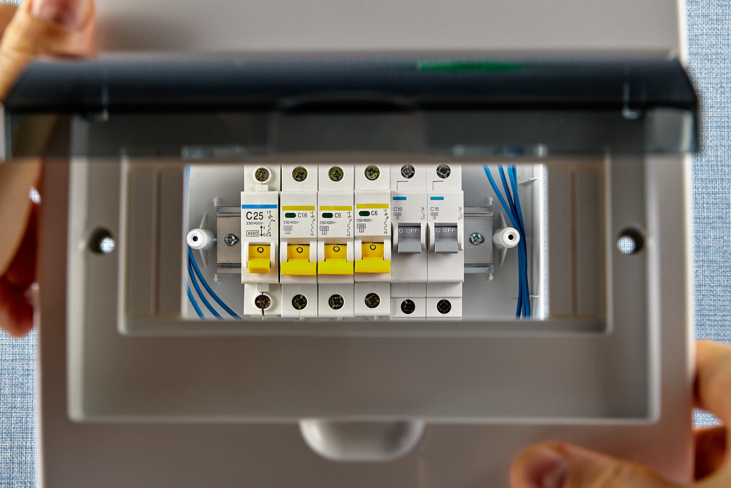 Hands are opening the cover of an electrical circuit breaker panel, conducting essential electrical safety checks. Inside, several circuit breakers with yellow switches and blue wiring are visible. The panel is mounted on a light blue wall.