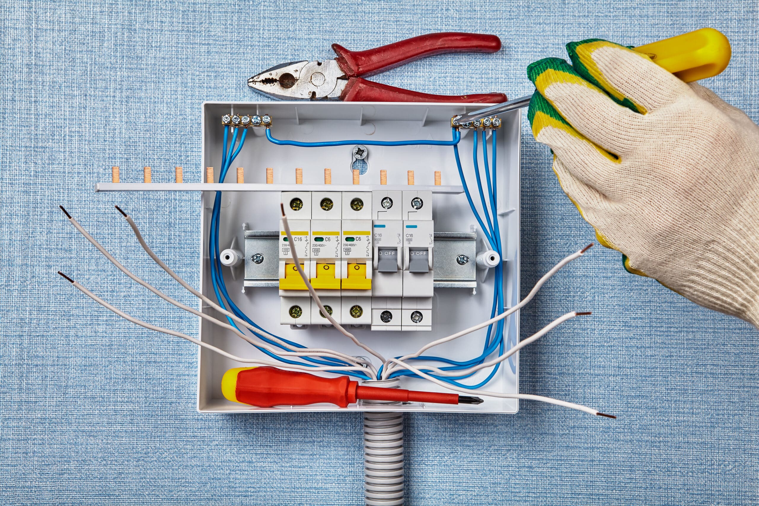 An open electrical junction box with exposed wires rests on a blue surface. A gloved hand holding a tool emphasizes the importance of electrical safety checks. Nearby, red-handled pliers and a screwdriver lie ready beside circuit breakers and colored wires.