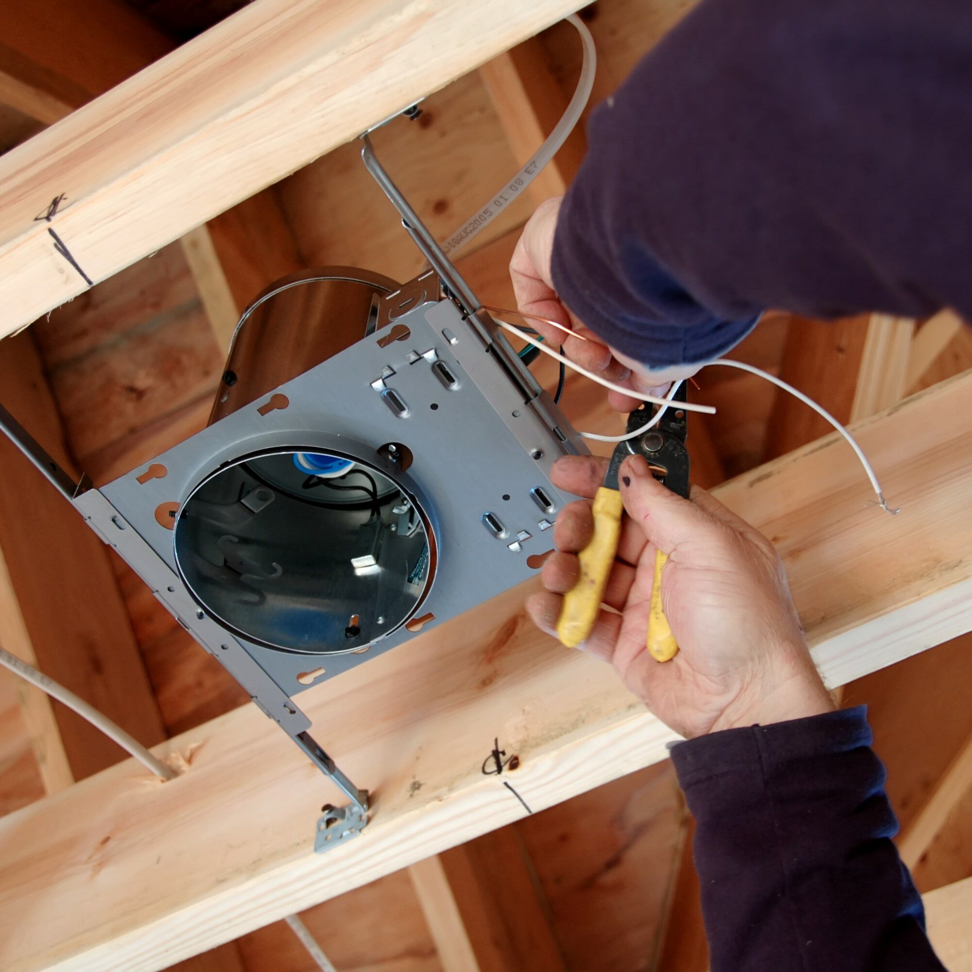 An electrician in a long-sleeve shirt installs a metal electrical fixture into a wooden ceiling frame. With precision, they use pliers to connect wires, ensuring thorough electrical safety checks are adhered to for optimal performance and security.