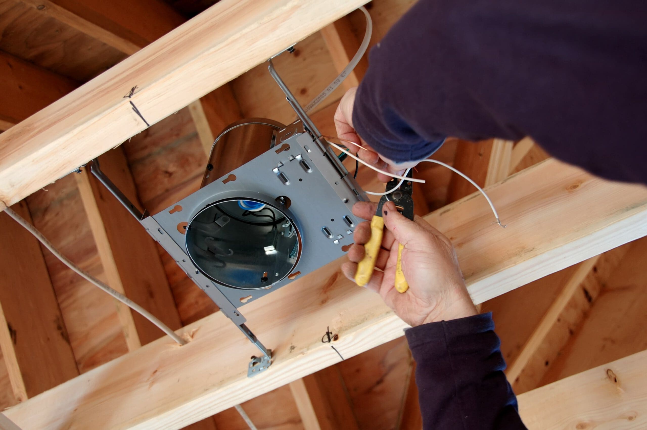 A skilled electrician is expertly wiring a recessed ceiling light fixture into the wooden framework. With yellow-handled tools in hand, they're diligently connecting wires as part of the electrical installation.