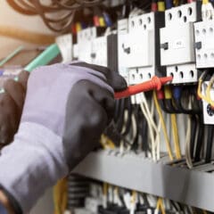 An electrician wearing gloves uses a multimeter to test voltage on a large circuit board with various wires and switches, expertly conducting electrical safety checks during maintenance work.