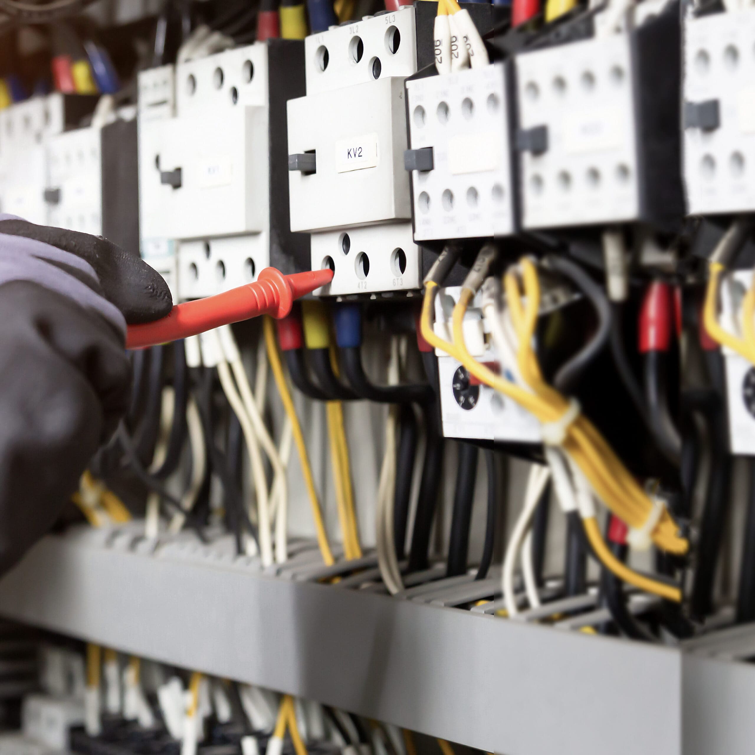 Close-up of an electrician using a screwdriver to repair wires in an electrical panel. The panel houses multiple circuit breakers, switches, and a complex network of colorful cables. The professional wears protective gloves, emphasizing the importance of electrical safety checks.