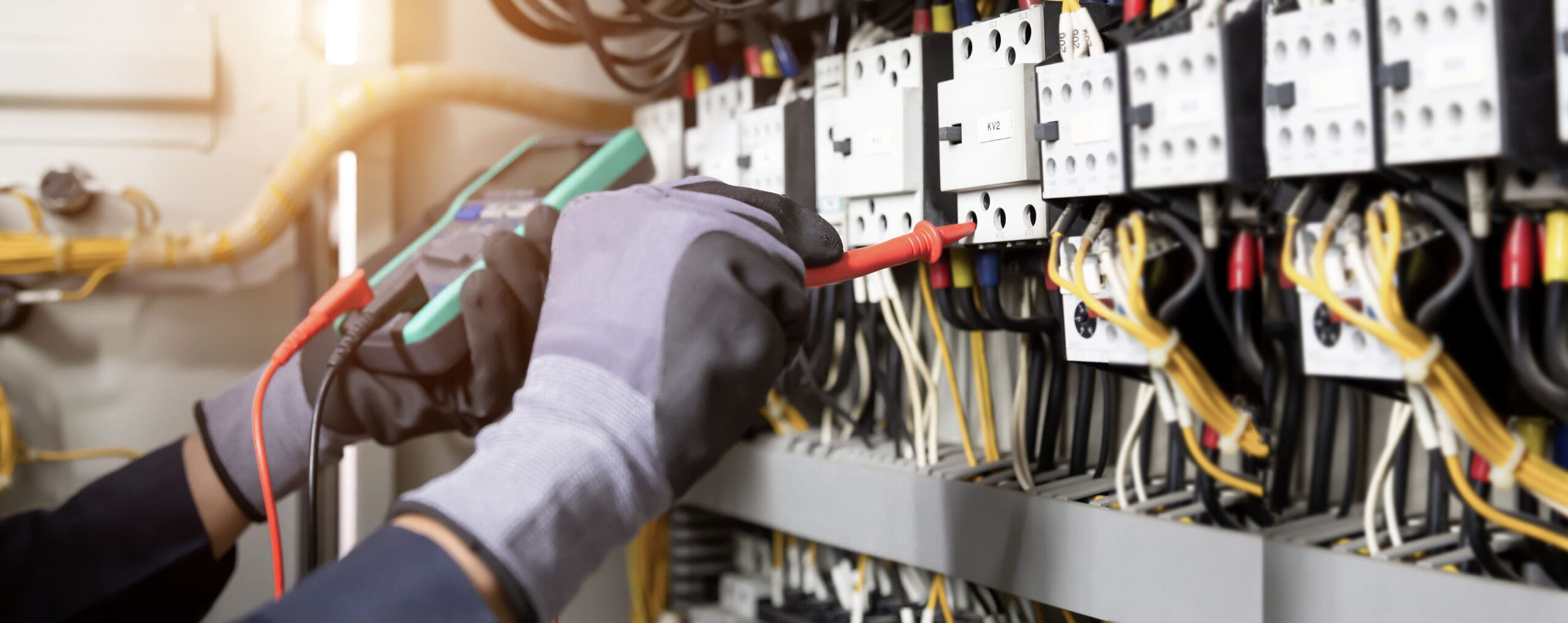 An electrician wearing gloves uses a multimeter to test voltage on a large circuit board with various wires and switches, expertly conducting electrical safety checks during maintenance work.