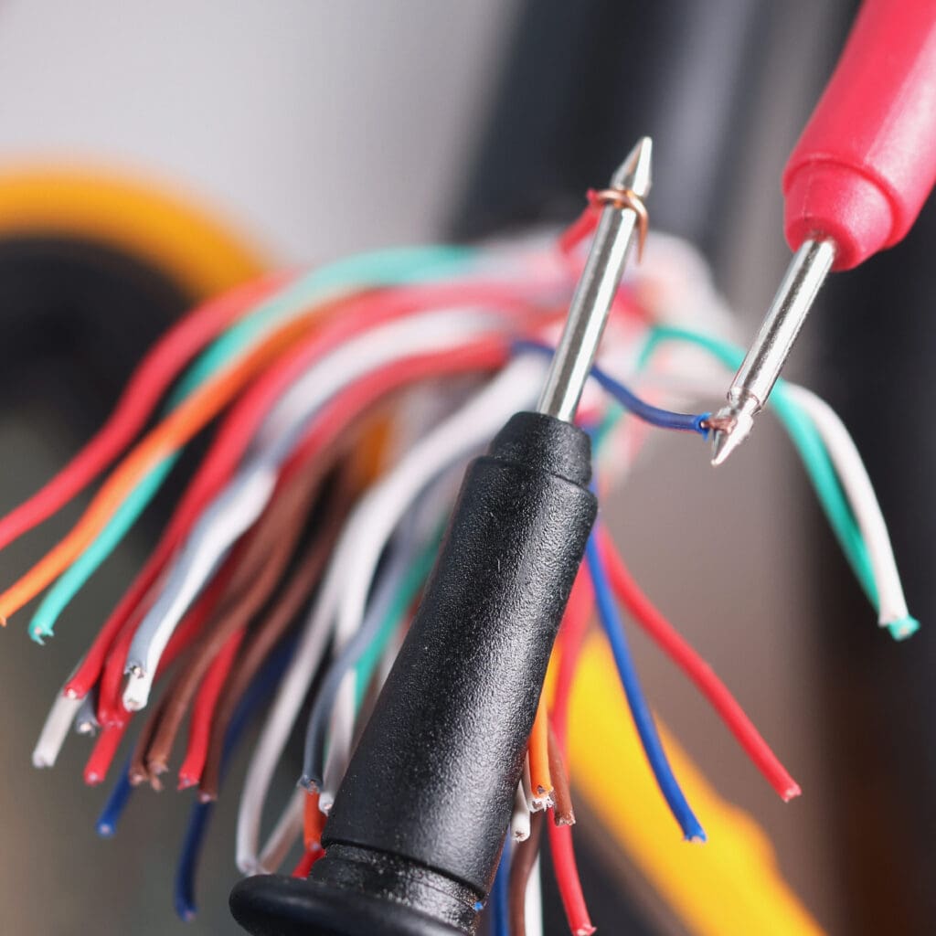 Close-up of multicolored electrical wires with two probes, one red and one black, making contact—ideal for fault finding. The background is slightly blurred, emphasizing the vivid colors and metallic tips crucial for efficient electrical safety checks.