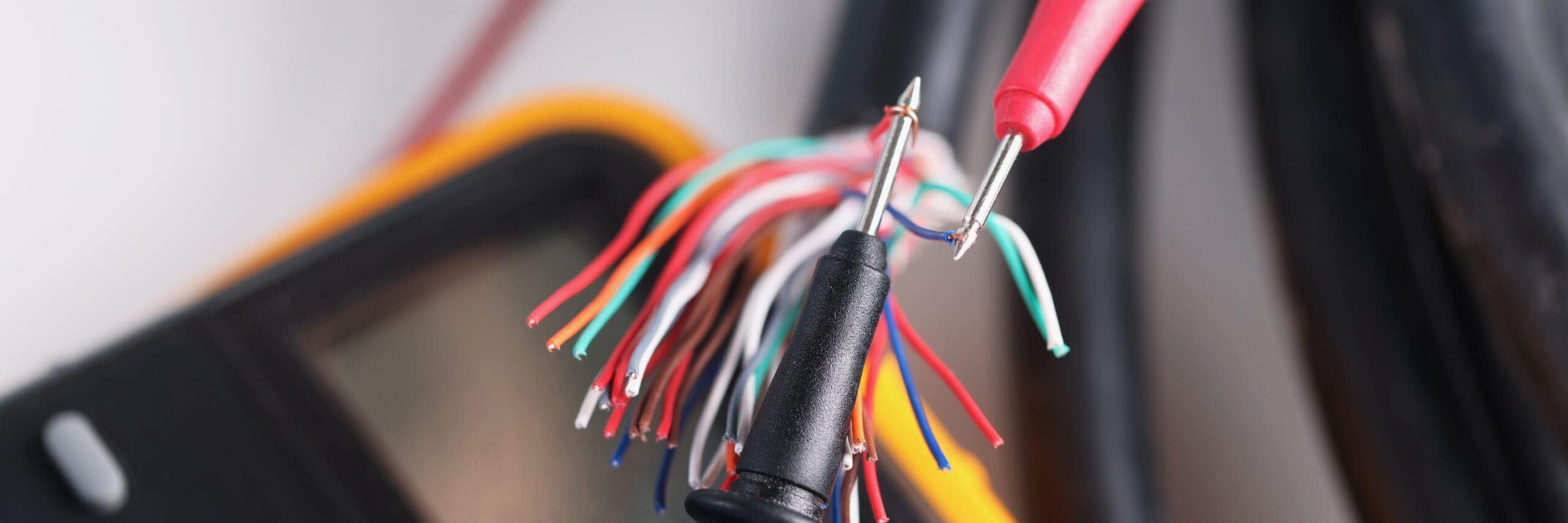 Close-up of multicolored wires with red and black probes from a multimeter, an essential tool for any electrician. The background is slightly blurred, showing part of the multimeter, suggesting electrical safety checks or diagnostics in preparation for a consumer unit upgrade.
