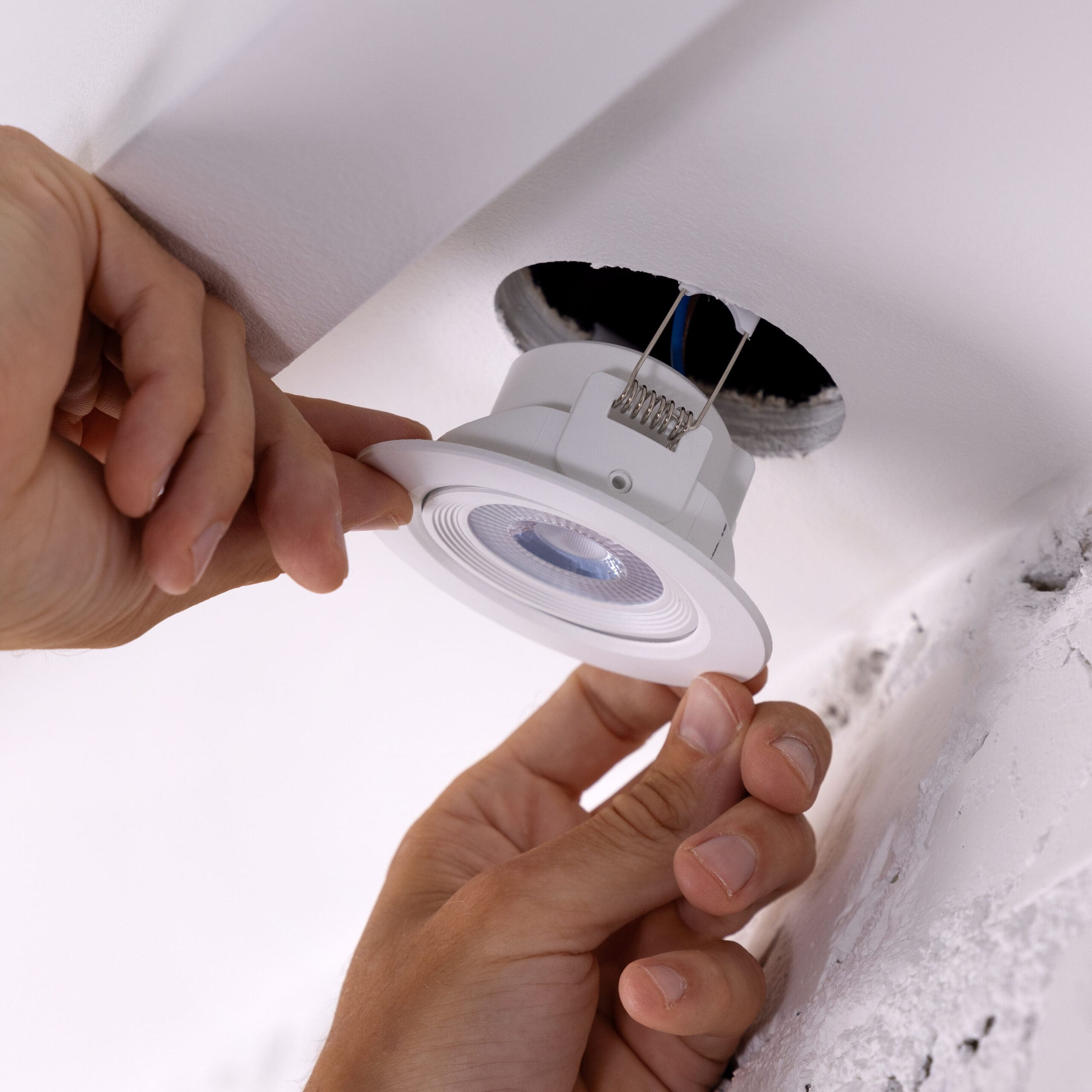 A person installs a white recessed ceiling light fixture. Their hands position the light near a hole in the ceiling, with wires visible. The ceiling is white with some slight imperfections around the hole.